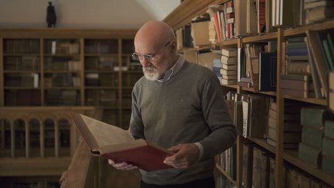 Mature male researcher in glasses standing near the bookshelves in the library, holding a book in his hands, looking to the camera and smiling. Indoors. Portrait.