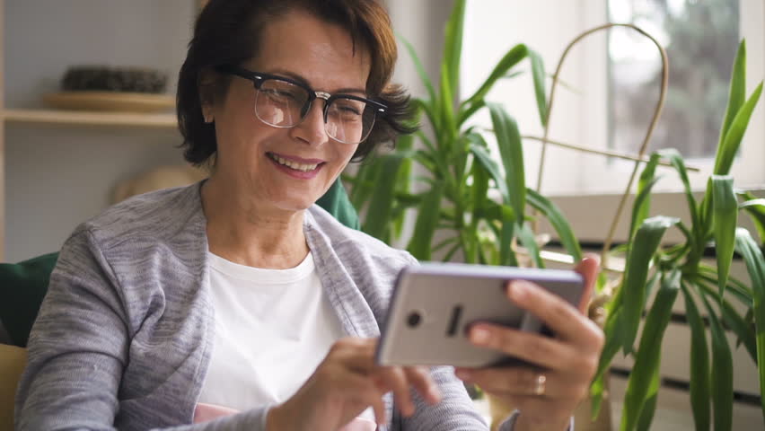 Happy mature woman in glasses smiling and waving her hand, having video call with family members on her brand new smartphone at home. Indoors. Portrait.