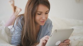Woman in blue shirt with brown hair lying on the bed with white sheets and surfin the internet on her brand-new tablet. Indoors. Portrait.