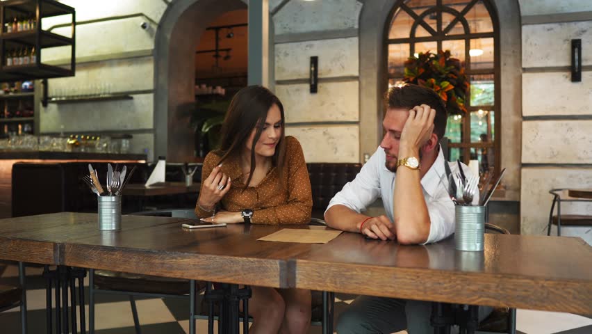 Nice couple are sitting together in a restaurant. She is drinking some tea and looking to a menu while her biyfriend is trying to pick food for them. Also he is giving food advices to her. | Shutterstock HD Video #1013794433