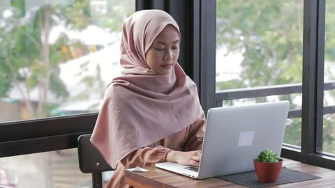 Portrait of smiling young muslim woman working on modern laptop in cafe. Attractive woman in hijab searching for something in internet. Edited and raw.