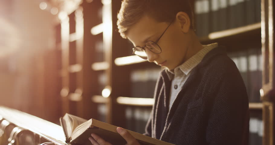 Portrait shot of the cute blond schoolboy in glasses reading a book attentively while standing in the library passage in sunlight. Quarantine
