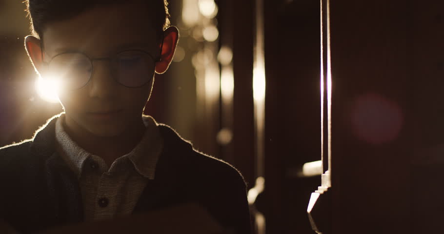 Portait of the cute teen boy in glasses reading a book while standing in the dark library in sunlight, then closing a book and sneezing because of the dust.