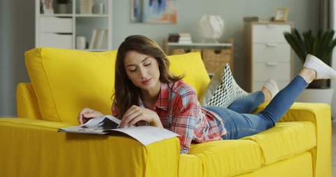 Pretty Caucasian young woman lying on the yellow couch, flipping pages and reading fashion magazine at home.