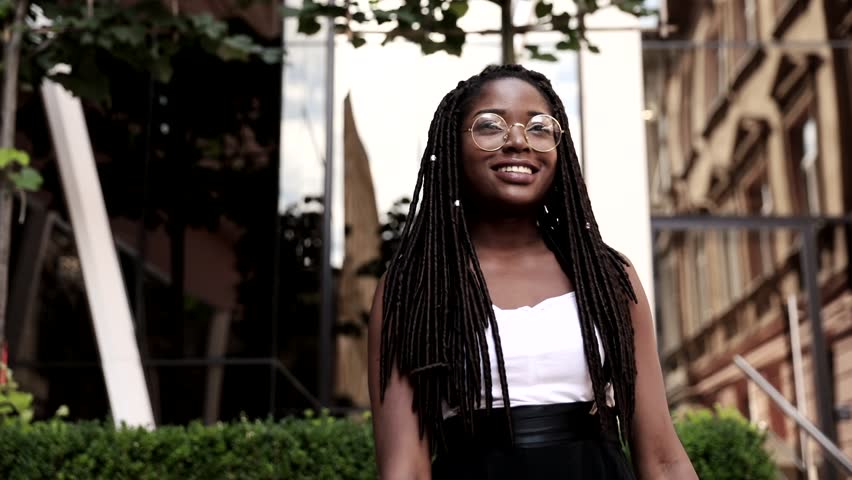 Portrait of Young Afro-american Girl Walking in the City. Happily Smiling Woman with Braids. Wearing Round Glasses.