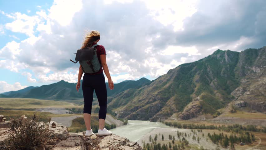A traveler with a backpack is standing on the edge of a cliff in the mountains and raises her hands up. back view