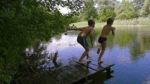 Two boys jump together from pier to the river in the forest, slow motion