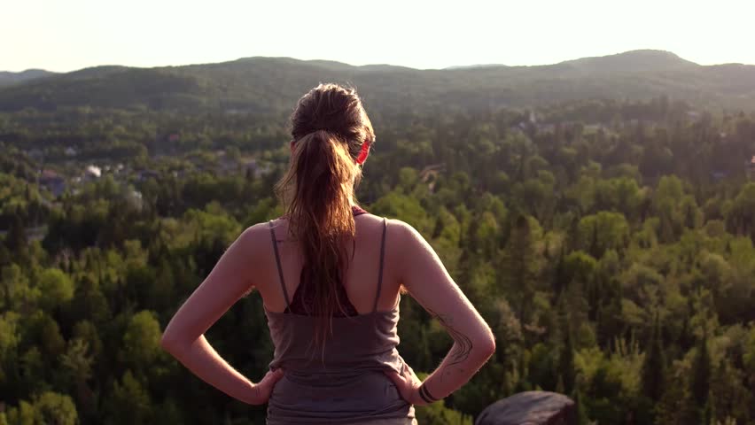 Girl standing up with arms resting at her waist while looking at the horizon on the top of a mountain with rock cliffs at sunset - Travelling up from behind her Royalty-Free Stock Footage #1013832980