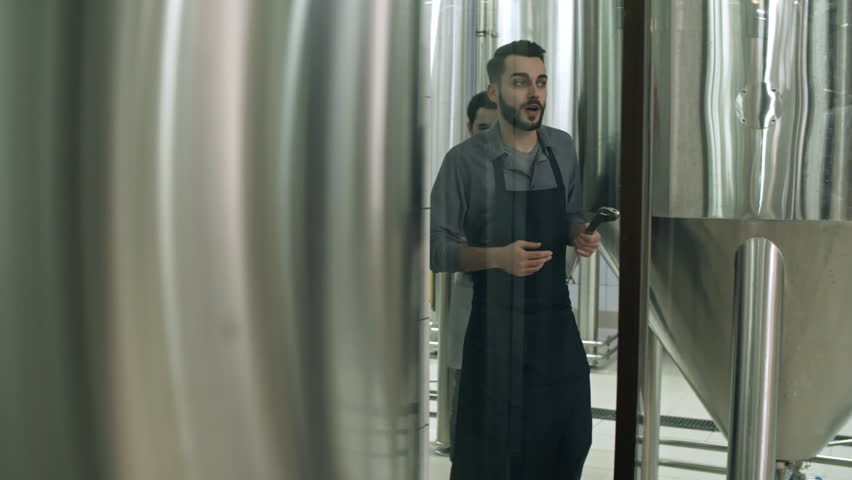 View through the glass wall of brewery technician in apron walking and talking to colleague in lab coat while walking in beer plant through steel vessels