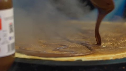 Slow motion vendor man cooking crepes on a metal griddle in Paris street. Close-up of pancake with chocolate dessert. Delicious fast food. A hand is making crepe outdoors. Cheff cook a tasty crepe-Dan