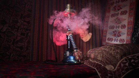 Hookah hot coals on shisha bowl making clouds of steam at Arabian interior. Oriental ornament on the carpet. Stylish oriental shisha in dark with backlight. Slider shot. Empty space