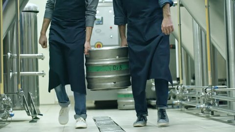 Low section of two brewery workers in aprons carrying together beer keg and walking towards the camera