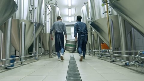 Close-up shot of two brewery workers lifting up beer keg together and walking away from the camera through brewhouse