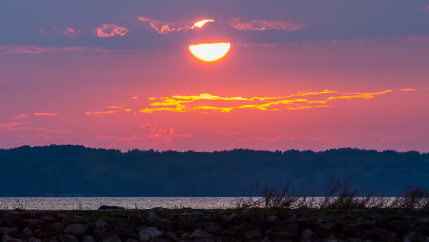 Timelapse of very orange sunset on small calm lake with moving clouds in background