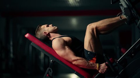 A muscular man performs exercises on a sports training apparatus for leg muscles in a dark gym, lifting weights
