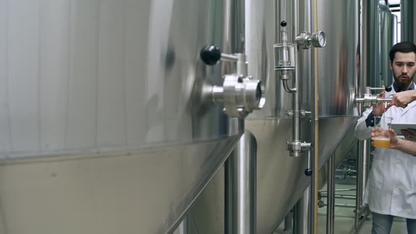 Tracking shot of brewery inspector in lab coat using digital tablet and talking to colleague while he pouring freshly brewed beer from vessel into jug in brewery