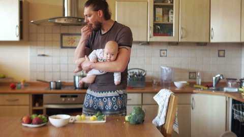 A young modern father cooks food in the kitchen, holding a three-month-old baby in his arms. Single parent, father's day. A man has a stylish beard and a ponytail on the back of his head