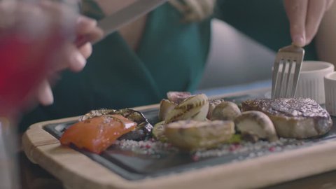 The food blurred concept: attractive blonde lady eating delicious steak in restaurant slow motion. Only hands, woman holding fork and knife and tasting meat with grill vegetables on the wooden plate