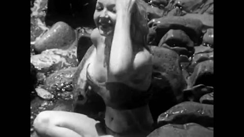 CIRCA 1950s - Pin-up girl Adele Dolman tries to sit on the rocks of a beach while the waves come in, but her bikini top slips a few times.
