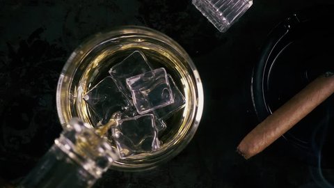 Slow motion pour whiskey into a glass with ice cubes and a smoking cigar in an ashtray top view