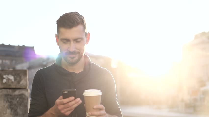 Happily Smiling Attractive Man Walking down the Street. Typing on his Smartphone with Interest. Enjoying the Evening Walk. Drinking Delicious Coffe. Crowded City on the Background. Royalty-Free Stock Footage #1013880362