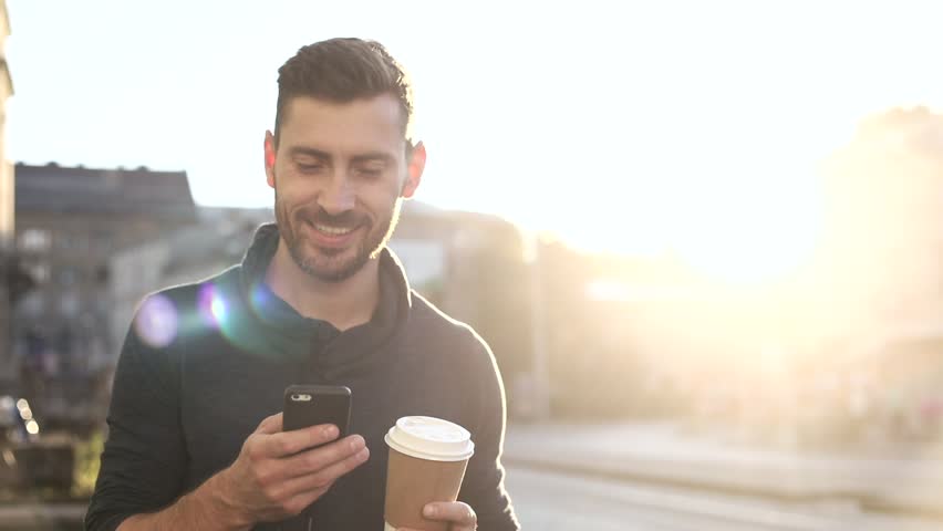 Happily Smiling Attractive Man Walking down the Street. Typing on his Smartphone with Interest. Enjoying the Evening Walk. Drinking Delicious Coffe. Crowded City on the Background. | Shutterstock HD Video #1013880362