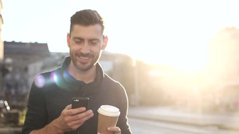 Happily Smiling Attractive Man Walking down the Street. Typing on his Smartphone with Interest. Enjoying the Evening Walk. Drinking Delicious Coffe. Crowded City on the Background.