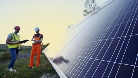 Male workers standing in uniform and helmets near solar panels outdoors in soft focus