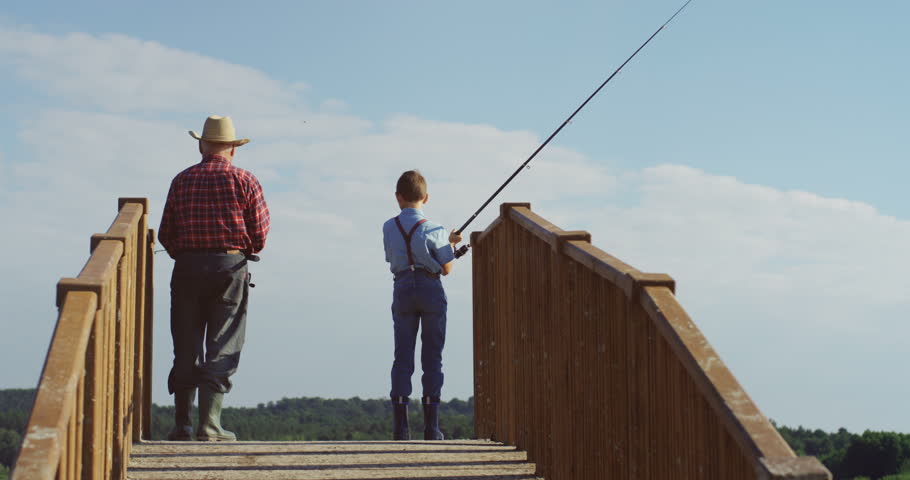Rear of the old grandfather fishing together with his little grandson while standing on the wooden bridge and holding rods in the water.