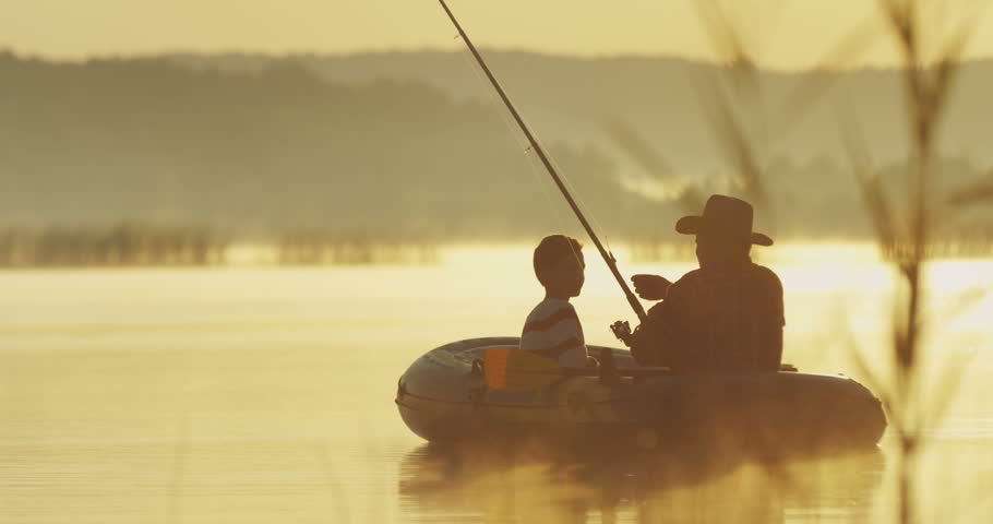 Old angler in a hat with a grandson sitting in the boat in the cane on the lake at the dawn with a rod while fishing. Outside. Royalty-Free Stock Footage #1013887865