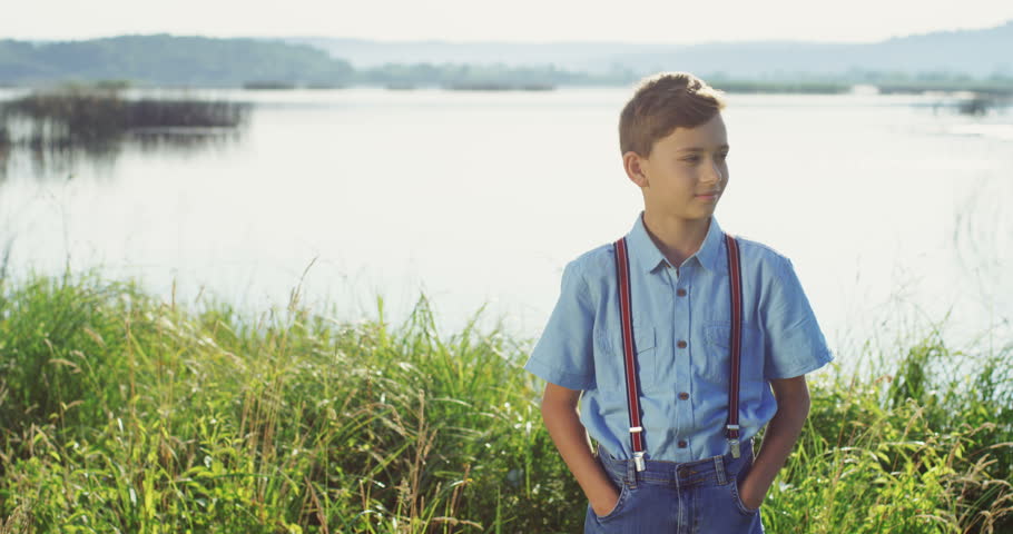 Portrait of the cute small teen boy standing on the big lake shore and lookin to the camera. Outdoor.
