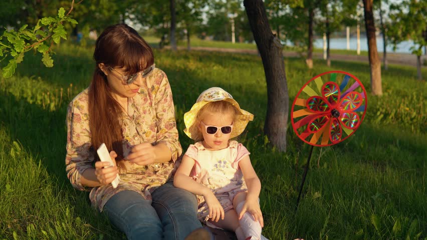 Mother and daughter in sunglasses have fun playing together on the grass under the young oaks at sunset. Young woman and little girl dancing listening to melodies in the smartphone app.