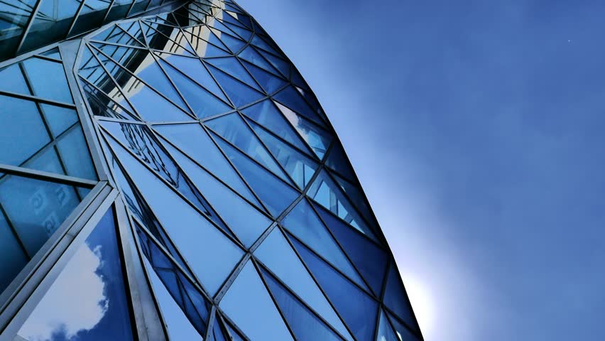 Time-lapse of a tall building with a glass angular panels. And the background of moving clouds during the day. Modern architecture. Blue colors and sunlight from behind the edge of the facade.