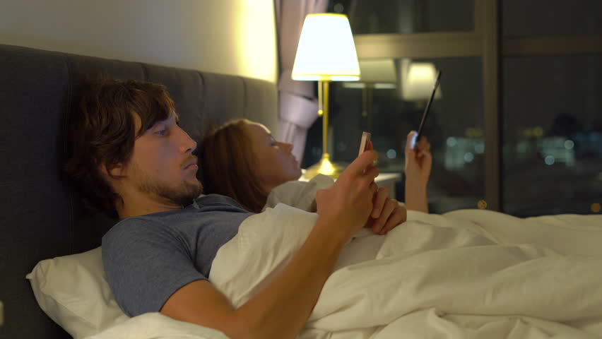 man and woman lying in bed each with smartphone and tablet pc. woman turns off the lamp and and goes to sleep while man not. problems in marriage and intersexual relationship problems and mobile