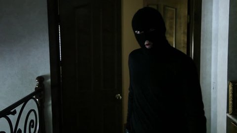 Wide shot of a masked intruder in black as he opens a door and creeps through the doorway looking around, then walks past camera.