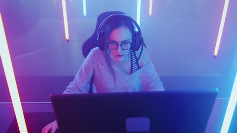 High Angle Shot of the Beautiful Friendly Pro Gamer Girl Playing in Online Video Game and Streaming it, Wearing Headset Talks with Her Fans and Team into Headphones Microphone. Shot on RED EPIC-W 8K.