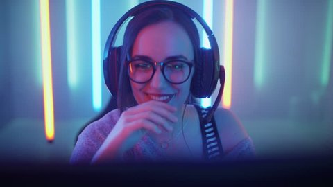 Beautiful Friendly Pro Gamer Girl Does Video Game Gameplaystream, Wearing Headset Talks with Her Fans and Team into Headphones Microphone. Background Cool Neon Retro Colors. Shot on RED EPIC-W 8K.