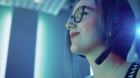 Low Angle Portrait Shot of the Beautiful Pro Gamer Girl Playing in Online Video Game, Cute Geek Girl in Glasses, talks with Team Players through Microphone. Neon Colored Room. Shot on RED EPIC-W 8K