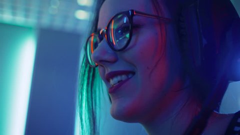 Low Angle Portrait Shot of the Beautiful Pro Gamer Girl Playing in Online Video Game, Cute Geek Girl in Glasses, talks with Team Players through Microphone. Neon Colored Room. Shot on RED EPIC-W 8K.