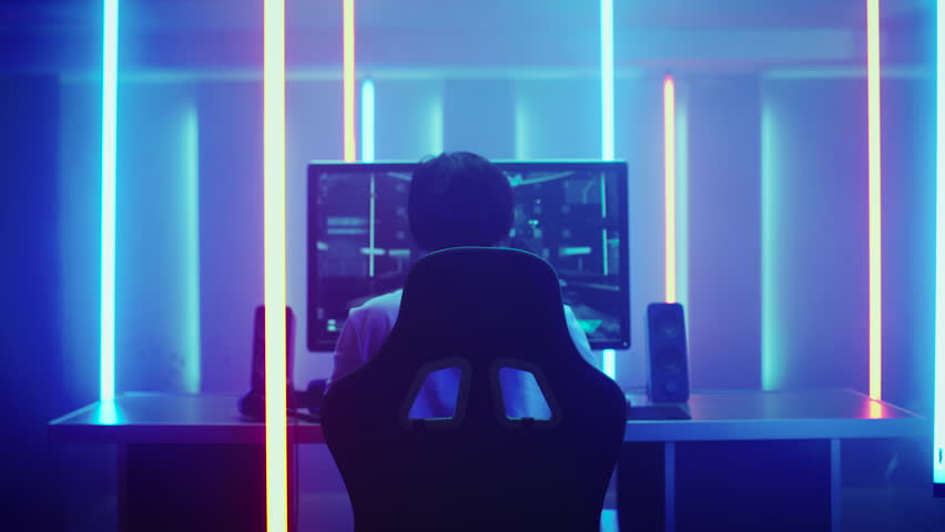 Back View Shot of the Professional Gamer Playing in First-Person Shooter Online Video Game on His Personal Computer. Room Lit by Neon Lights in Retro Arcade Style.  Shot on RED EPIC-W 8K Helium Camera