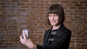 Young woman with short haircut having video call through her cell phone, isolated on brick background