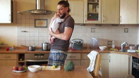An attractive father cooks soup in the kitchen holding his little son in his arms. Dad on maternity leave