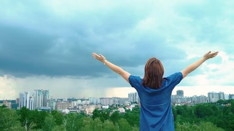 The girl raises her hands to the sky, standing on a hill away from the city, the view from the back