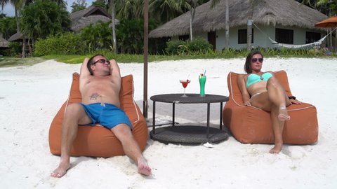 A man and a woman lie on a padded stools on the beach and drink cocktails