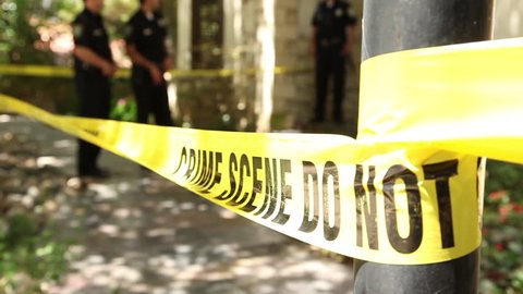 Closeup shot of police tape at a crime scene, with cops guarding the perimeter of a home in the background as a detective enters and exits through the scene.