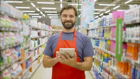 Handsome smiling supermarket employee with beard using digital tablet standing among shelves In supermarket