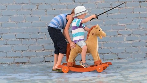 Amusing child rolling his younger brother on toy rocking horse with plastic sword. Boy dreams of battles, victories and adventures.Concept of moral education, patriotism, hero, super. Children happy