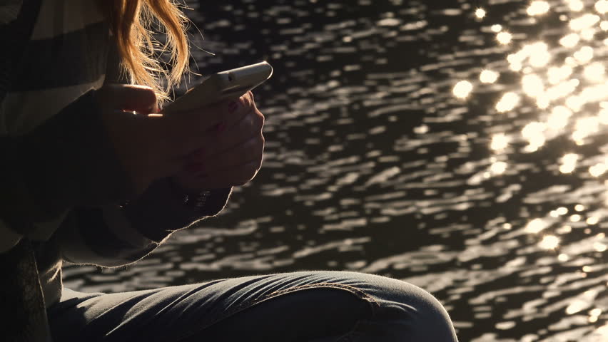 relaxed woman typing on her smartphone near the river at sunset