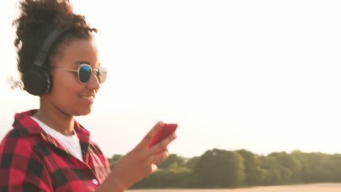 Tracking video clip of beautiful mixed race African American girl teenager young woman wearing a red and black shirt and blue sunglasses walking listening to music on wireless headphones at sunset 