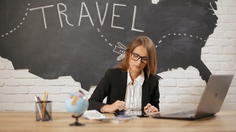 Intelligent female travel agent registrating tourists online, using tablet and laptop to fill in passport information, wearing white blouse and black suit on typical working day